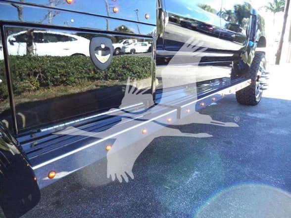 Heavy haul truck side view showing black exterior, chrome steps, and large tire. Image includes reflection of surroundings and a faint watermark silhouette of a running dog.