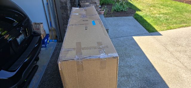 Boxed truck part shipment placed on driveway beside a dark vehicle with a garage in the background.