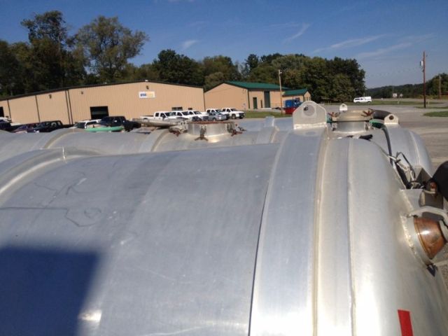Tank trailer top with manway lids and fittings; background shows industrial buildings and parked trucks.