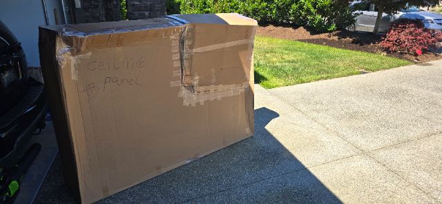 Cardboard box on driveway with taped seams and handwritten text on the side. The text reads 'Ceylon Panel' or similar scribble. Shadowed driveway, green lawn in background.