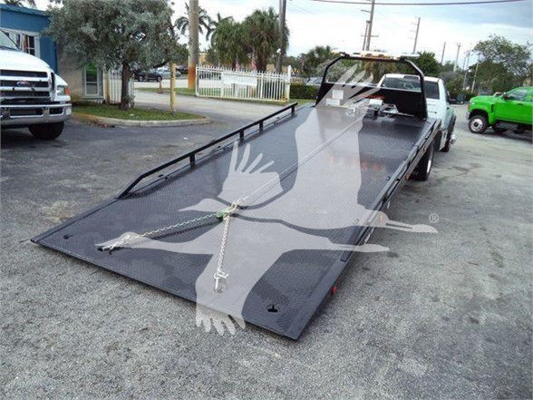 Ram 5500 tow truck with a flatbed trailer in a dealership lot. Front cab visible in background, raised steel ramp bed with chains securing center, white truck behind.