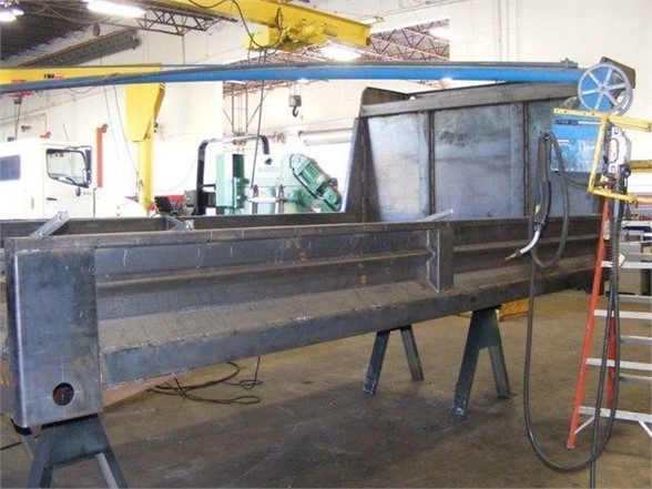 Industrial workshop showing a long rectangular metal dump body component on sawhorses with a blue overhead crane and a wheel mechanism at the end.