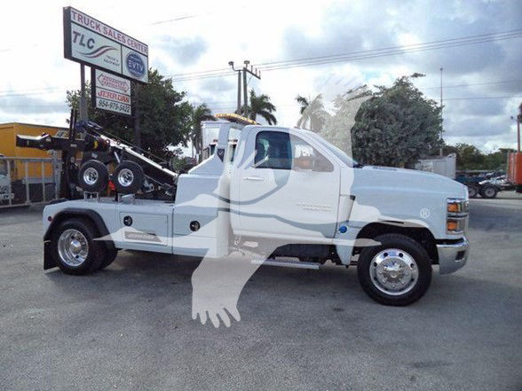 Truck with tow body and wheel-lift equipment. Visible text includes TLC Truck Sales Center sign in background. No clear model-specific labels readable on the truck.