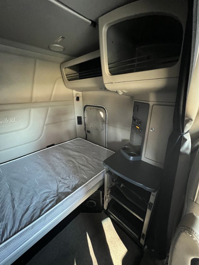Peterbilt 579 truck sleeper cab interior showing single bunk bed, upper storage shelf, and small desk area with controls.