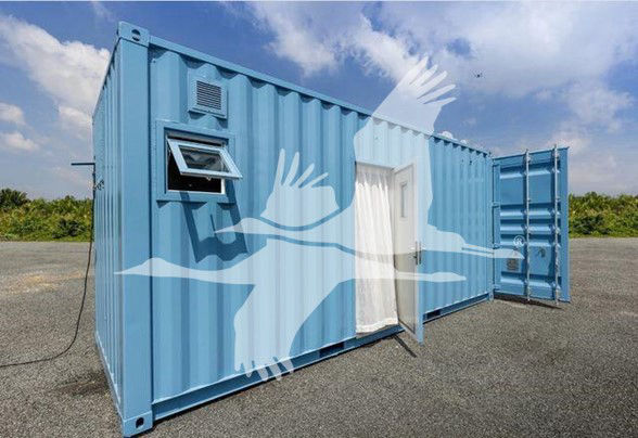 Blue modified shipping container trailer with a side window and a door, open rear ramp, and a vent near the front. Appears to be a converted trailer or storage unit.