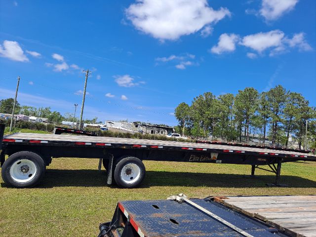 Flatbed trailer in a field with trees and a blue sky. Visible text on the trailer reads 'Elite Parts' along the side. The image shows a long black flatbed with dual rear axles and white wheels.