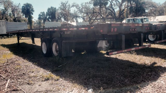 Flatbed trailer showing extended trailer frame with dual rear axles and reflectors. Visible tires and undercarriage; some branding plates and metal railing along sides.