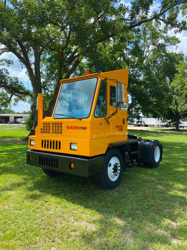 Yellow Ottawa yard spotter 4x2 in a grassy lot with green trees. Cab shows 'KALMAR OTTAWA' branding, large front grille, black bumper, white wheels, two exhaust stacks near cab.