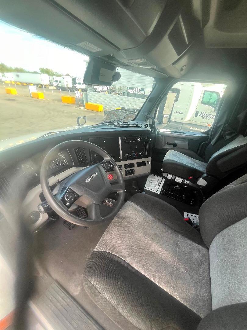 Truck cab interior of a 2021 Freightliner Cascadia 126: steering wheel, center console, dashboard with infotainment screen, driver seat, passenger seat, and gear shifter.