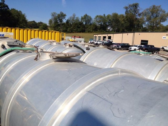 Large aluminum vacuum tank trailers with rounded cylindrical modules, fittings and hoses on top. Background shows a yellow container, vehicles, and a beige building.