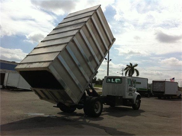 16 ft dump truck body mounted on a tilt cab truck, metal box tilted upward for dumping, with a white truck cab and industrial lot in background.