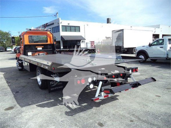 Flatbed tow truck with orange cab and long aluminum bed, rear view showing towing equipment and lights.