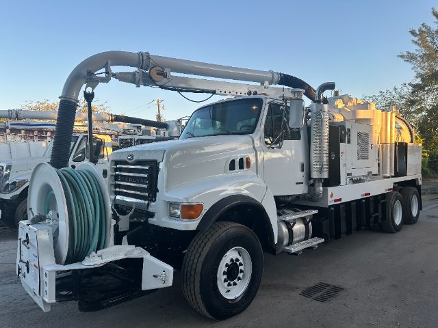 Vacuum truck body on a white Sterling L8500 with large hose reel and crew cab, side storage compartments, and elevated exhaust in the rear.