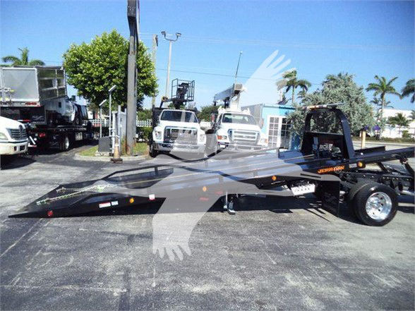 Flatbed tow rig on a dealership lot with Ram 5500 truck and other vehicles in the background. Visible text on the trailer includes some orange and white markings and a logo silhouette overlain in the image.