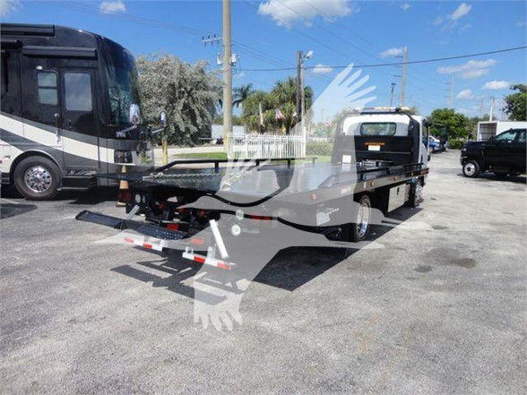 Isuzu NRR truck with a flatbed tow bed, rear view shows trailer hitch and hydraulic system; background includes another large motorhome, utility poles, and blue sky.
