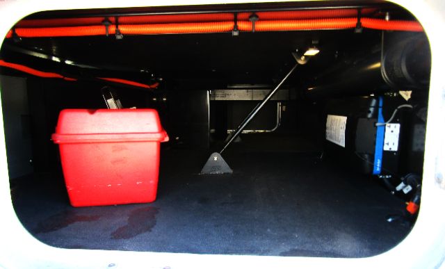 Underbelly storage area of a Kenworth T680 cab and chassis showing a red storage box, black floor, support prop, and a blue strap with a rectangular white label.
