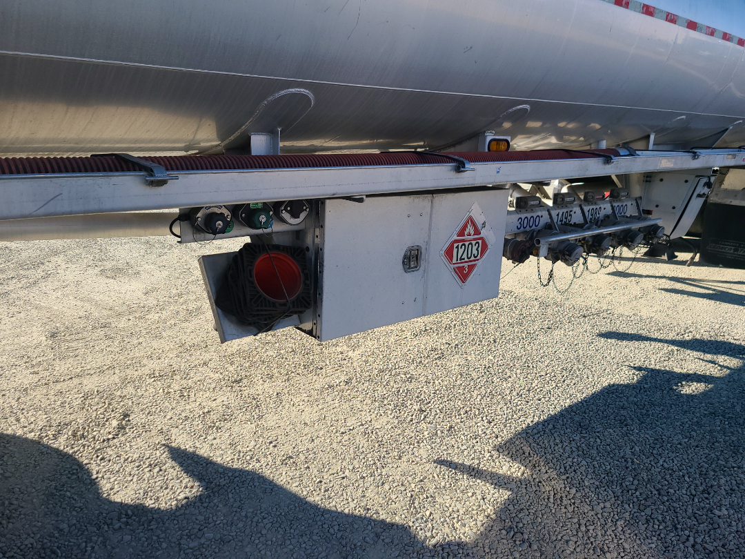 Beall semi-trailer with a side-mounted pump module, warning placard 1203, and multiple valve connections along its undercarriage.