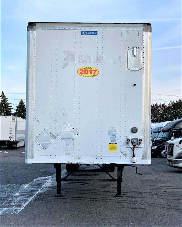 Stoughton dry van trailer rear view showing raised landing gear, rear doors removed badge, and stickers: Stoughton logo at top center, 2017 model sticker, various safety placards and wiring near lower right corner.