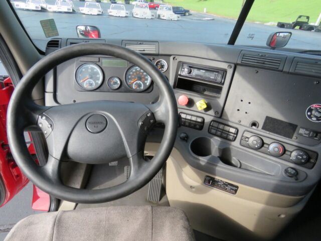 Truck cab interior showing steering wheel with Freightliner badge, instrument cluster, center console with controls, and radio in dashboard.