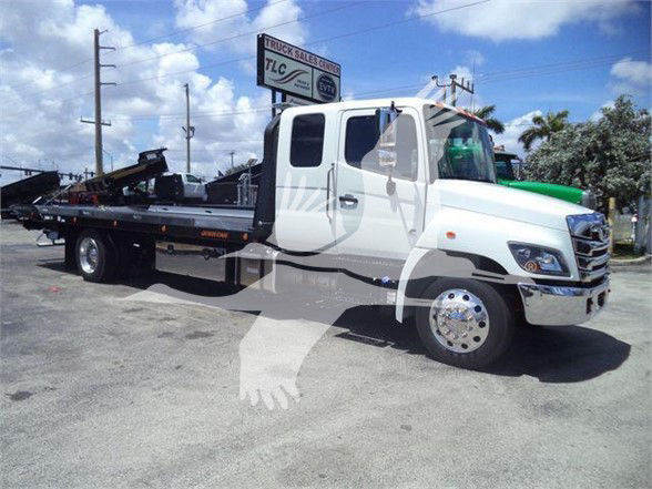 Truck with a flatbed tow setup showing front cab and long trailer bed. Visible text on signage in background: 'TLG' or similar. No detailed engine labels visible.