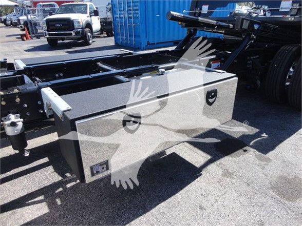 Mack MD6 truck chassis with a large aluminum tool/storage box mounted on the frame, visible fuel or hydraulic lines, and a blue container in the background.