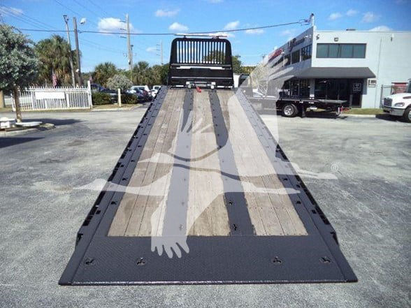 Flatbed tow truck with a long wooden deck and metal frame, viewed from the rear approaching the platform.