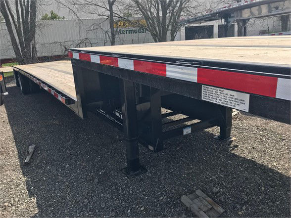 Flatbed trailer with black frame and wooden deck, Gilbert red-and-white safety reflectors along the edge, label on side with specifications, support legs visible.