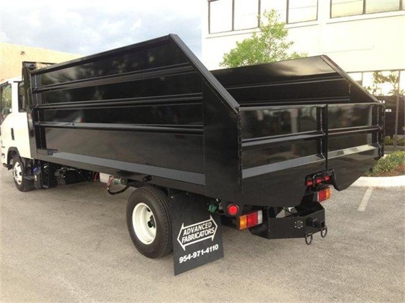 Truck bed dump body with black metal sides and tailgate on a white cab truck; Advanced Fabricators sign on the ground near rear wheel.