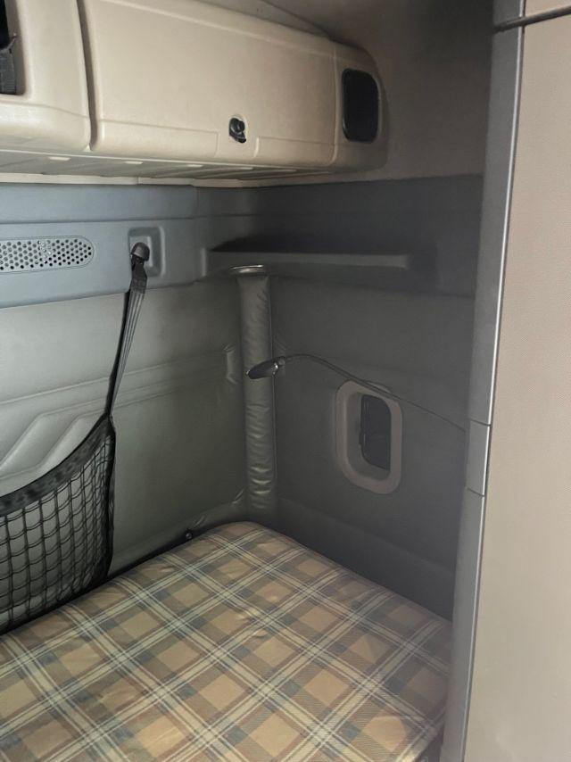 Freightliner Cascadia sleeper cab interior showing bunk area with plaid mattress, side storage, and overhead compartment.