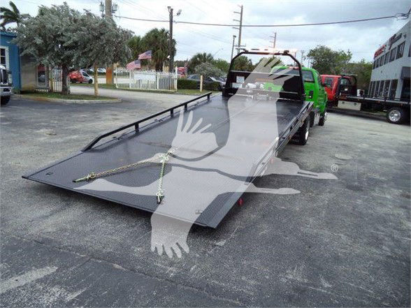 Flatbed tow truck with a large black loading ramp extended, tie-down chains across the deck, green tow truck in background, dealership lot.