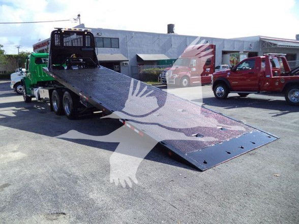 Tow truck with a long metal ramp on a flatbed trailer, Freightliner M2 106 in background, red pickup and green tow truck visible.