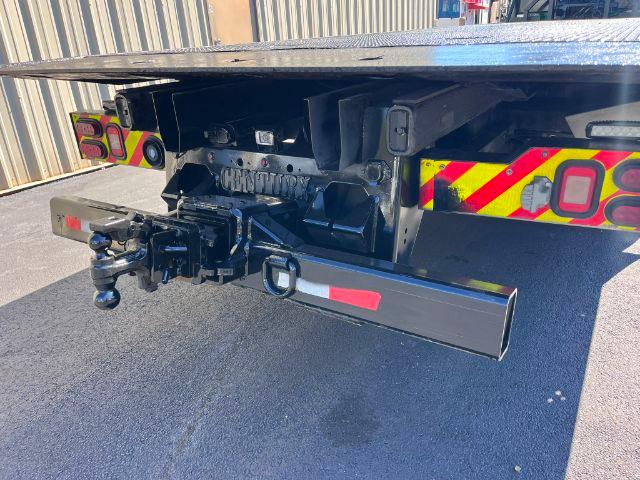 Peterbilt 348 rollback truck rear with tow hitch, hydraulic components, and a flatbed extension visible. There are reflective safety chevrons on the sides and a black metal bumper/undercarriage assembly with hooks and a hitch receiver.