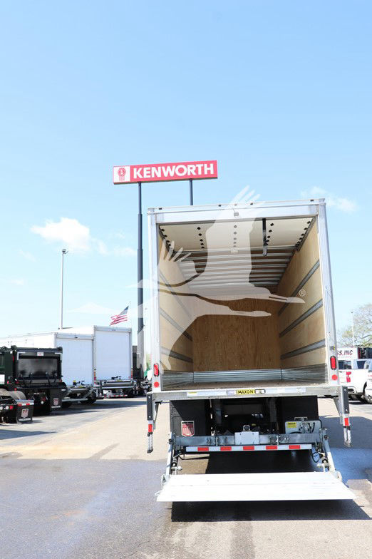 Back of a truck with an empty cargo box in a dealership lot. A Kenworth sign is visible in the background.