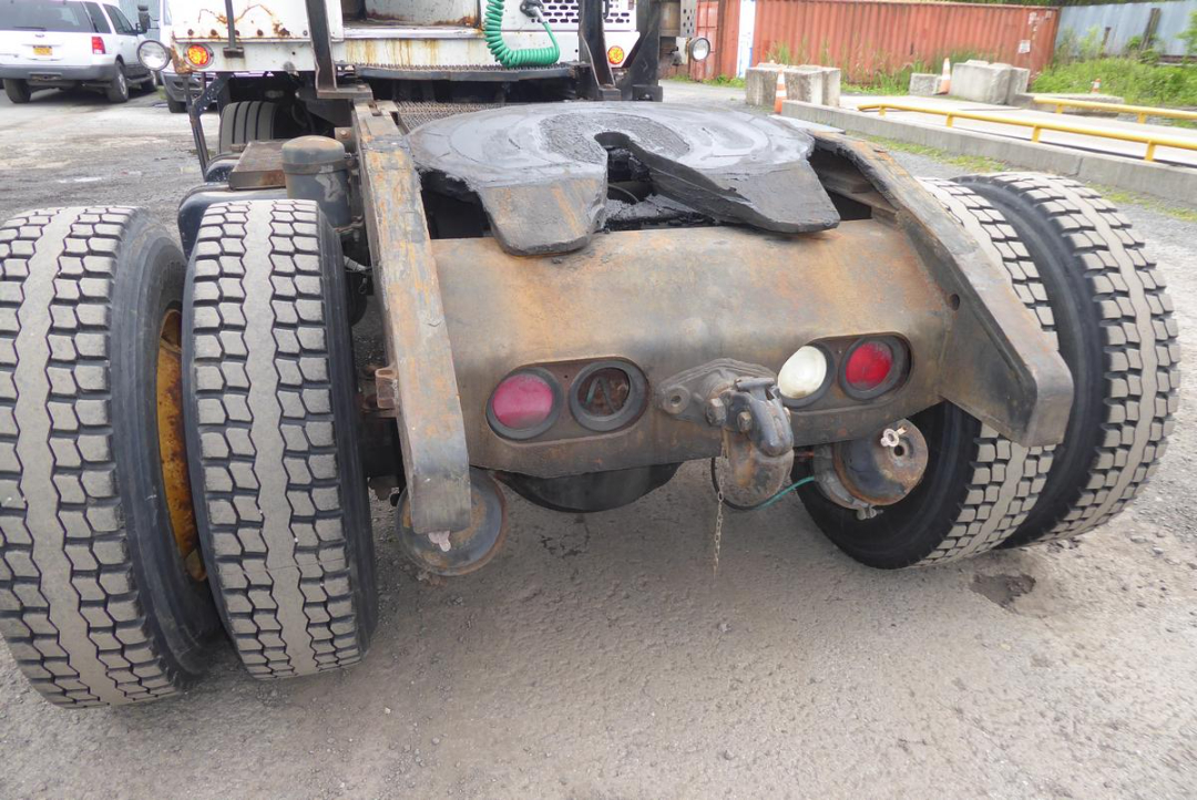 Rear view of a heavy truck chassis showing twin rear axles with dual tires, rusted frame, and hitch assembly with red and white taillights.