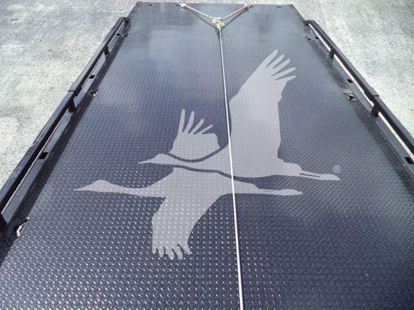 Flatbed truck trailer with diamond plate surface and a crane or winch strap at the center, featuring a large white crane logo across the deck.