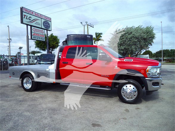 Ram 5500 SLT flatbed truck side view with red cab and silver flatbed. Visible text on building sign in background includes TLC Truck Sales Center and other smaller signs.