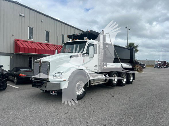 Kenworth T880 dump truck in a parking lot, white cab, black dump body, rear wheels, stock number TJ219615LM notations on billboard in background.