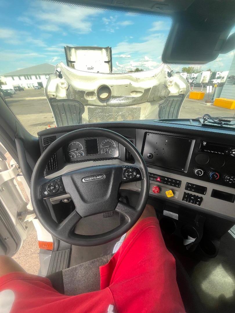 Interior of a 2021 Freightliner Cascadia 126 truck cab showing steering wheel, dashboard, and control panels with red shorts worn by occupant.