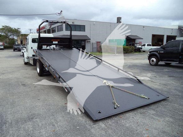Freightliner M2 106 tow truck with a long flatbed ramp down. Vehicle is white with a gray bed, parked in lot with building in background.