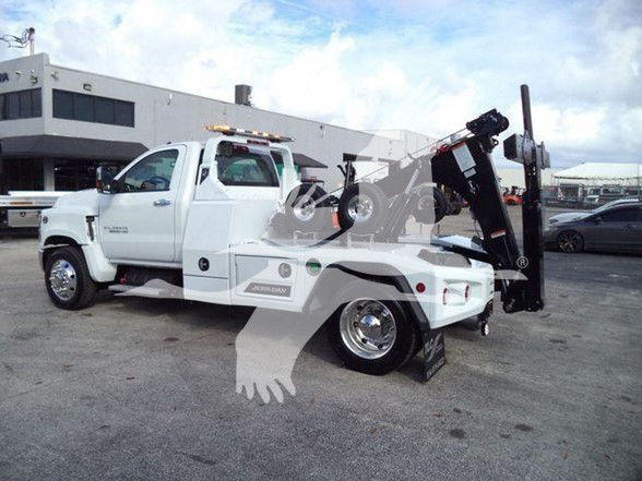 Tow truck body with a white Chevrolet Silverado 6500HD, front cab visible, hydraulic tow crane at rear, dealership lot in background.