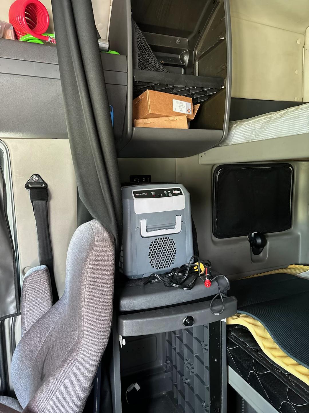 Truck cab interior showing a gray portable cooler device on a shelf, with cables and a small speaker grill, storage cubbies, and a bunk bed area.
