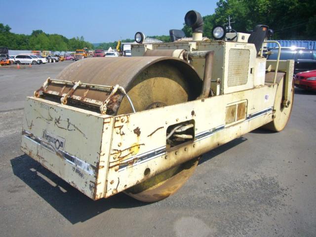 Tractor-style heavy roller compactor with worn white paint; large steel drum roller at front; operator seat and controls visible; side panels with rust and scratches.