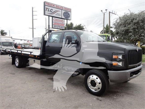 Truck in tow configuration with flatbed, cab front view, International CV model. Visible branding on truck and dealership sign behind.