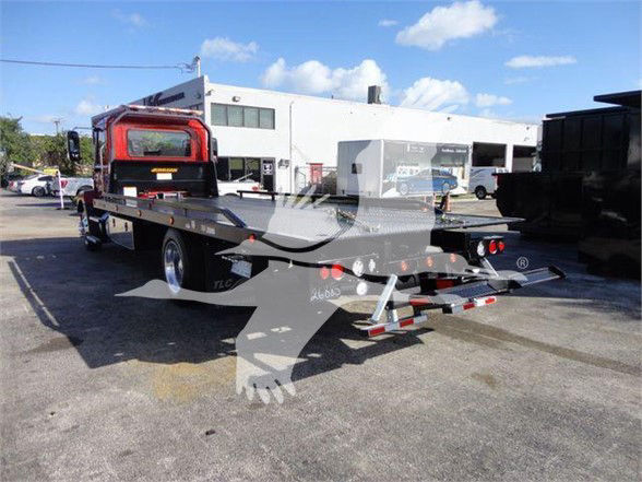 Flatbed tow truck with long flatbed, rear ramp, Mack logo watermark. Visible stickers and tail lights on rear, industrial buildings in background.