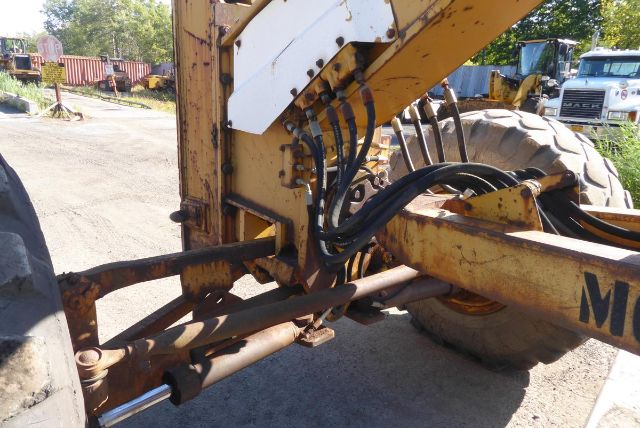 View of a Caterpillar 16G front hitch and hydraulic lines with rusty metal frame and large tires visible in background.