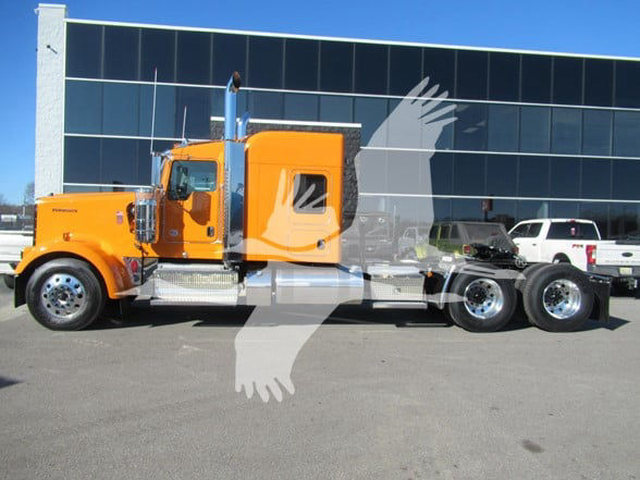 Kenworth W900L conventional sleeper truck in orange with chrome exhaust stacks and side steps, three rear axles, parked in front of a glass-front building.