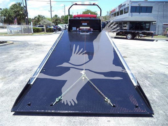Kenworth tow truck flatbed with black diamond plate; rear ramp extended; safety chains and hooks visible; faint Kenworth badge on cab; parking lot and building in background.