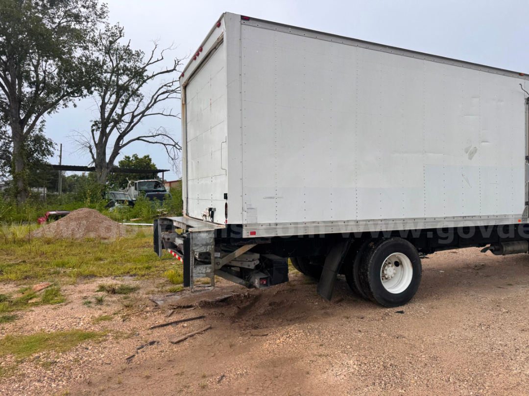 Box truck trailer with rear wheels visible and back of cab; white cargo box, black undercarriage, gravel ground.