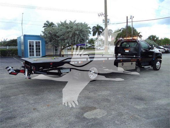 Chevrolet Silverado 6500HD tow truck with flatbed trailer and black pickup truck in a dealership lot.