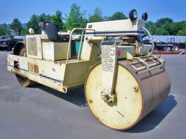 Tractor-drawn single drum roller, from side-left, with operator seat and control panel. Visible text on warning tag attached to front frame and small sticker on side panel.